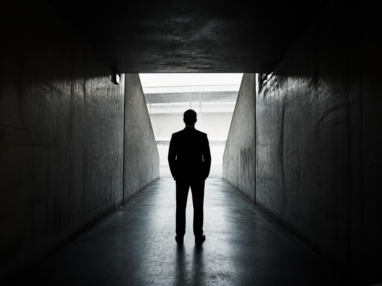 A person in a dark suit standing alone in an empty, backlit stadium tunnel